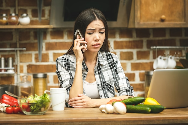 Mujer subrayada asiática hermosa joven con la computadora portátil en cocina. Usando el teléfono. Trabajando a casa. En estres.