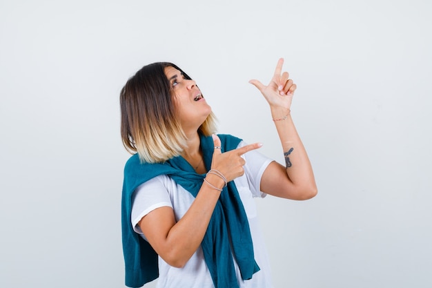 Mujer con suéter atado apuntando hacia arriba y hacia la derecha en camiseta blanca y mirando sorprendido, vista frontal.