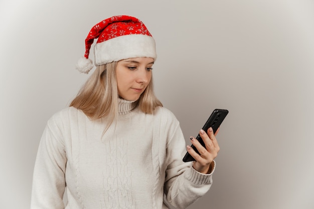 Mujer con un suéter blanco y gorro de Papá Noel con un teléfono en sus manos sobre un fondo gris aislado