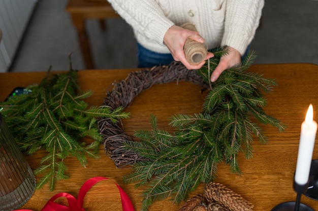 Una mujer con un suéter blanco hace una corona navideña de coníferas en una mesa de madera con velas Taller de coronas navideñas El tema de las vacaciones de Año Nuevo es un lugar para su diseño