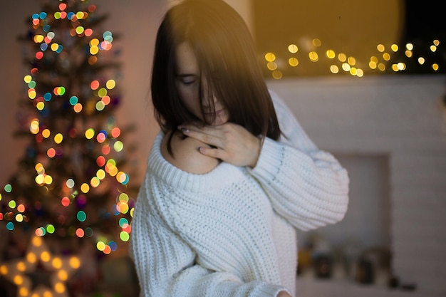 Una mujer en suéter blanco sonriendo y delante del árbol de Navidad y la chimenea