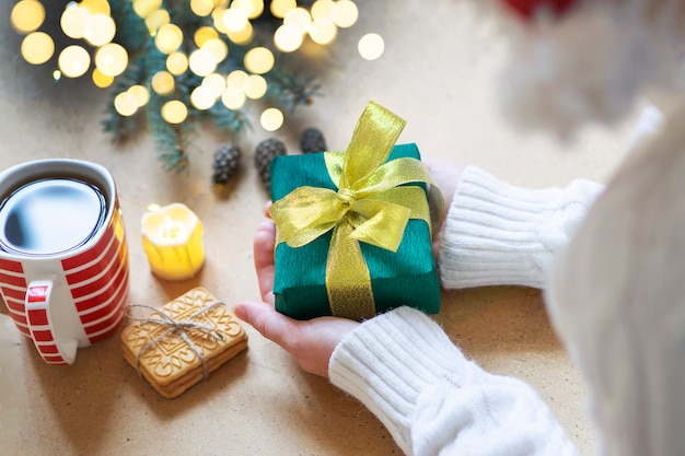 Una mujer con un suéter blanco sostiene un regalo de Navidad sobre un fondo de mesa de madera. Foto atmosférica con luces bokeh. Enfoque selectivo. Concepto de regalos para Navidad y año nuevo.