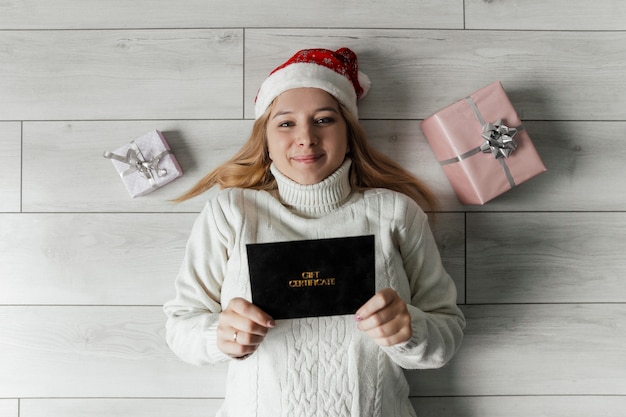 una mujer con un suéter blanco yace en el suelo y tiene un certificado de regalo en sus manos