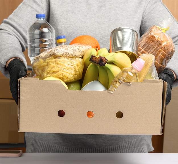 Mujer en suéter gris y guantes negros sosteniendo una caja de cartón con comestibles, concepto de asistencia y voluntariado, entrega de alimentos