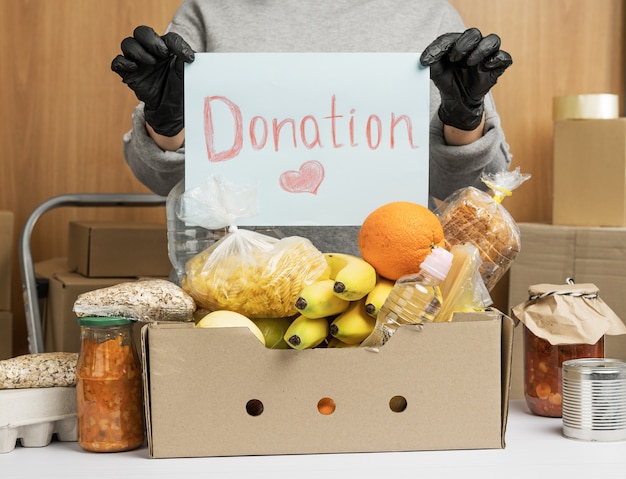 Mujer con un suéter gris y guantes sostiene una hoja de papel con la inscripción donación, en la mesa hay una caja de cartón con comida y frutas. Concepto de voluntariado