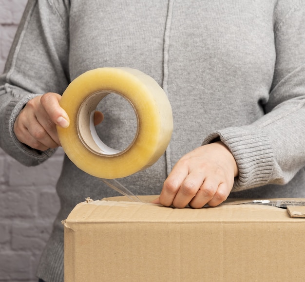 Foto mujer con un suéter gris sostiene un rollo de cinta adhesiva y empaca cajas de cartón marrón. concepto en movimiento