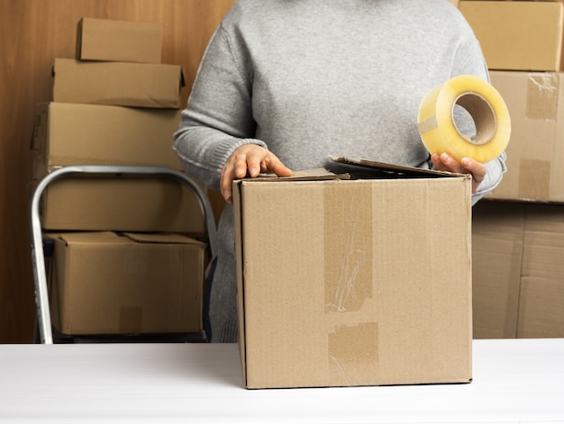 Mujer con un suéter gris sostiene un rollo de cinta adhesiva y empaca cajas de cartón marrón sobre una mesa blanca, detrás de una pila de cajas. Concepto en movimiento