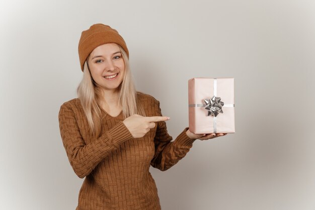 Mujer con un suéter marrón y un sombrero sosteniendo un regalo rosa en sus manos sobre un fondo gris