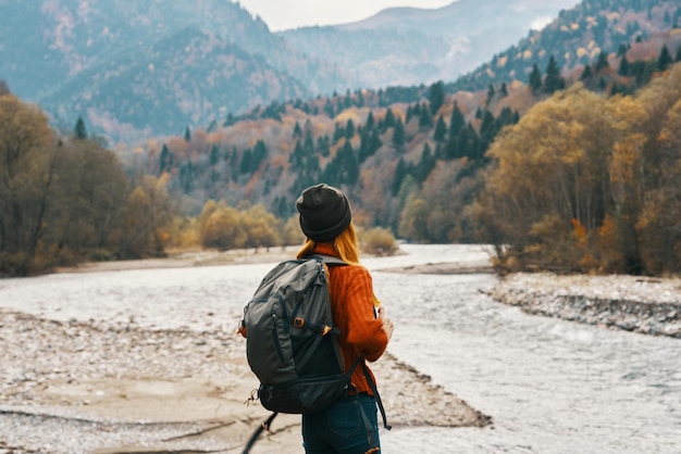 Mujer en suéter con mochila descansando en las montañas cerca del río en otoño en la naturaleza y modelo de árboles altos