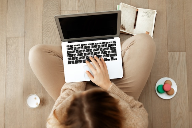 Mujer con un suéter de punto beige trabaja en la computadora sentada en el suelo