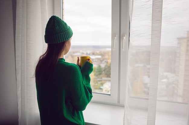 Mujer con un suéter de punto verde y un sombrero se encuentra en la ventana de la casa con una taza de café en otoño y mira a lo lejos en el bosque