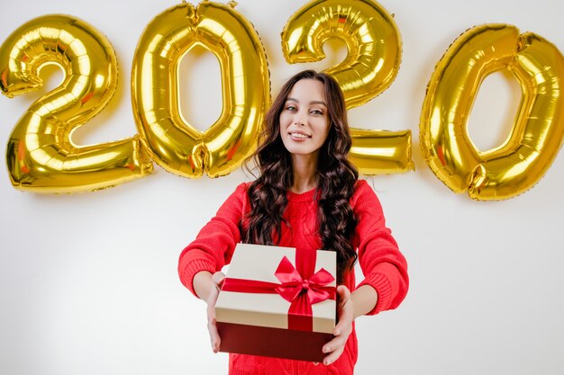 Mujer en suéter rojo abriendo regalo de Navidad delante de 2020 globos de año nuevo