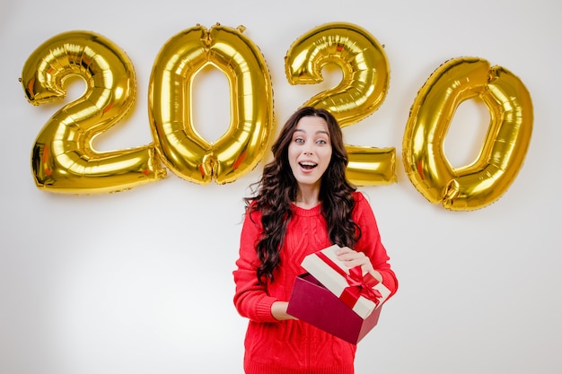 Mujer en suéter rojo abriendo regalo de Navidad delante de 2020 globos de año nuevo