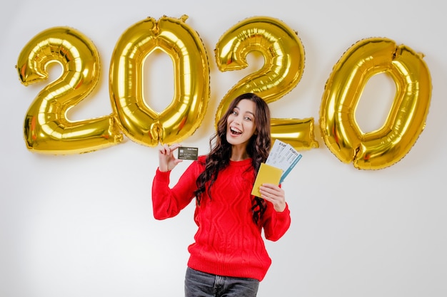Foto mujer en suéter rojo con plantilla de tarjeta de crédito y boletos de avión frente a 2020 globos de año nuevo