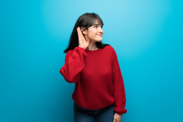 Mujer con suéter rojo sobre pared azul escuchando algo poniendo la mano en la oreja