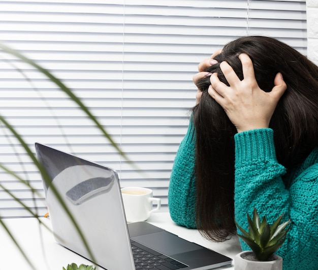 Una mujer con un suéter se sienta a la mesa con la cabeza entre las manos, estrés y dolor de cabeza por exceso de trabajo, fatiga. Una computadora portátil y una taza de café están sobre la mesa.