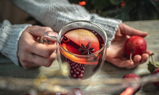 Mujer con un suéter sostiene una taza transparente con vino caliente especiado picante fragante decorado con frutas y especias. Concepto de ambiente festivo acogedor, año nuevo y humor navideño.