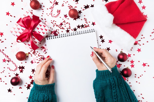 Mujer en suéter verde escribiendo lista de verificación de planes y sueños para el próximo año. Lista de deseos para Navidad. Lista de tareas para el nuevo año 2020 con decoración roja de vacaciones.