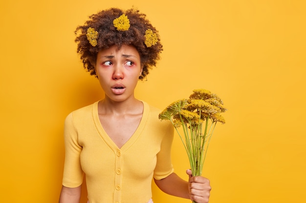 La mujer sufre de una reacción alérgica a la planta con flores tiene un aspecto poco saludable enrojecimiento de los ojos tiene flores silvestres aisladas sobre amarillo