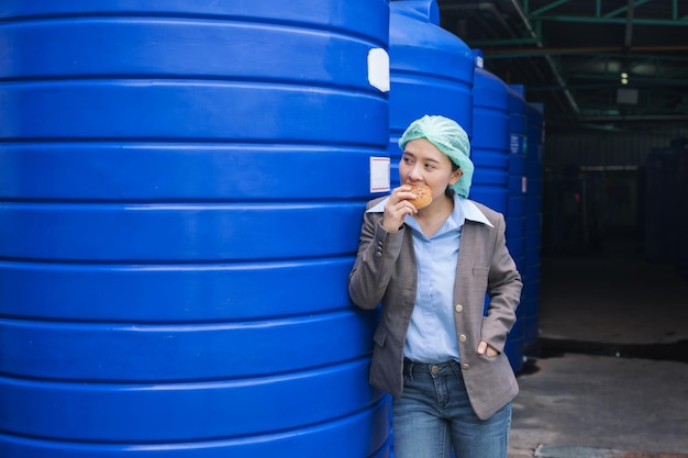 Mujer supervisora asiática de pie comiendo pan con prisa durante el descanso del almuerzo en el almacén de tanques de agua en la fábrica de procesamiento