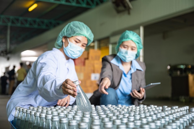 Mujer supervisora de inspectores asiáticos con un empleado auxiliar vestido con traje estéril comprobando el producto en stock de envases de bebidas de frutas en el almacén de la planta de procesamiento