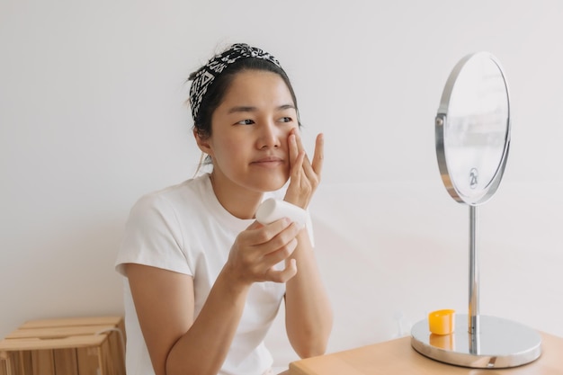 Foto mujer tailandesa asiática usando y aplicando crema hidratante solar frente al espejo mientras está sentada en