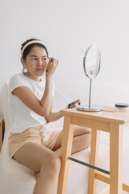 Foto mujer tailandesa asiática usando lápiz y dibujando cejas sentada en un sofá blanco frente al espejo en