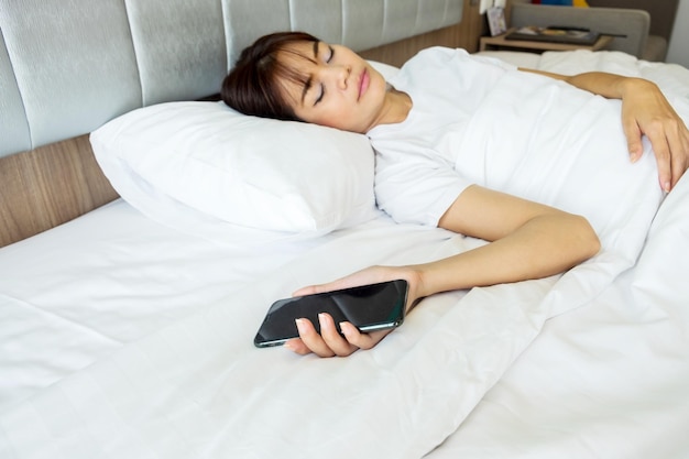 Una mujer tailandesa con una camiseta blanca se queda dormida mientras juega con su teléfono inteligente en una cama blanca en una habitación con luz que brilla a través de la ventana.