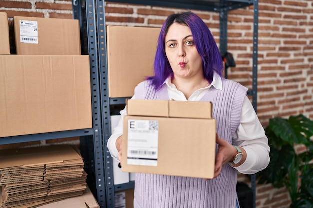 Foto mujer de talla grande con cabello morado que trabaja en una pequeña empresa de comercio electrónico sosteniendo cajas que hacen cara de pez con la boca y los ojos entrecerrados, loca y cómica.