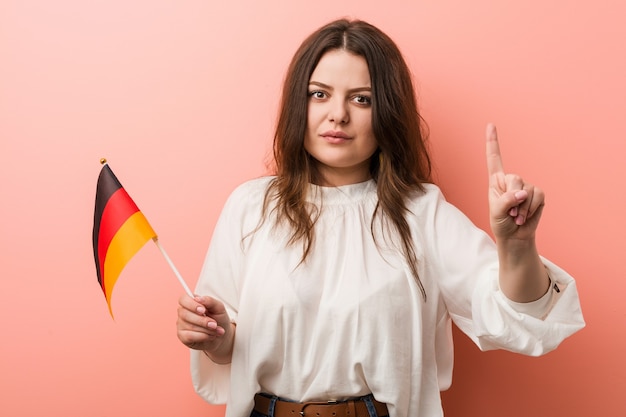 Mujer de talla grande con curvas joven sosteniendo una bandera de Alemania mostrando el número uno con el dedo.