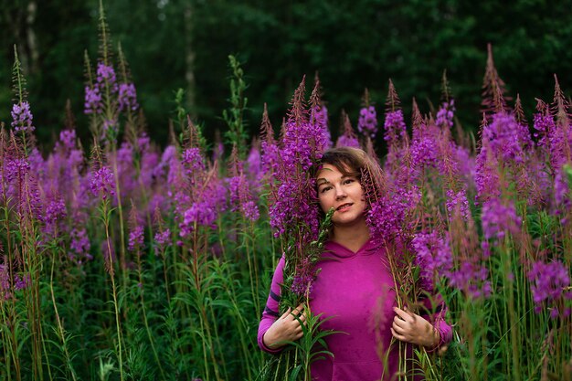 Mujer de talla grande en fucsia hoodie mirando a la cámara mientras está de pie en medio de flores brillantes en la pradera