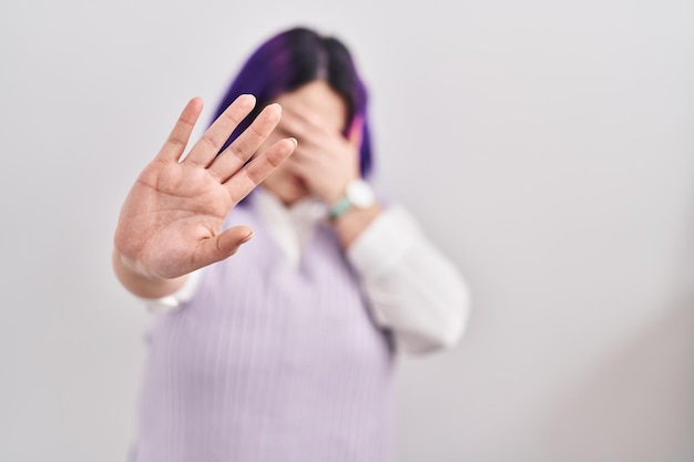 Mujer de talla grande con pelo morado sobre fondo blanco cubriendo los ojos con las manos y haciendo un gesto de alto con expresión de tristeza y miedo. concepto avergonzado y negativo.