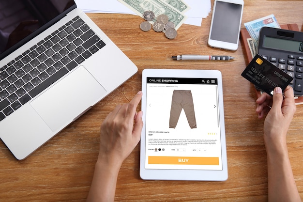 Mujer con tarjeta de crédito para comprar pantalones jogger marrones en el sitio web de comercio electrónico a través de una tableta con computadora portátil, teléfono inteligente y papelería de oficina en el escritorio de madera