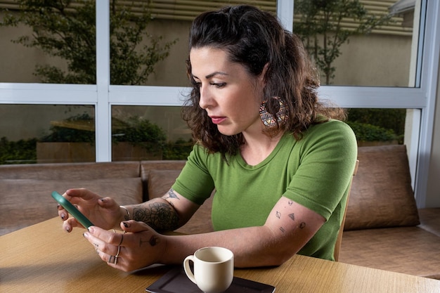 Mujer tatuada brasileña sentada junto a una taza de café e interactuando con la vista de su teléfono inteligente