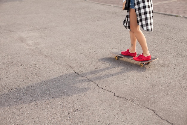 La mujer con tatuaje en camisa larga está patinando sobre un patín sobre el fondo del asfalto.