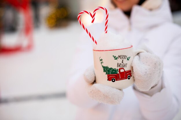 Mujer con taza con bastón de caramelo de nieve e inscripción Merry and Bright en sus manos al aire libre con ropa de abrigo en el mercado festivo de invierno Guirnaldas de luces de hadas decoradas ciudad de nieve para Navidad de año nuevo