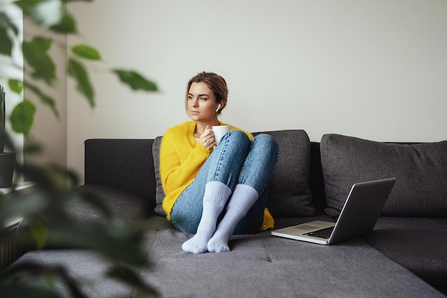 Mujer con una taza de café caliente sentada en el sofá y usando una computadora portátil en casa