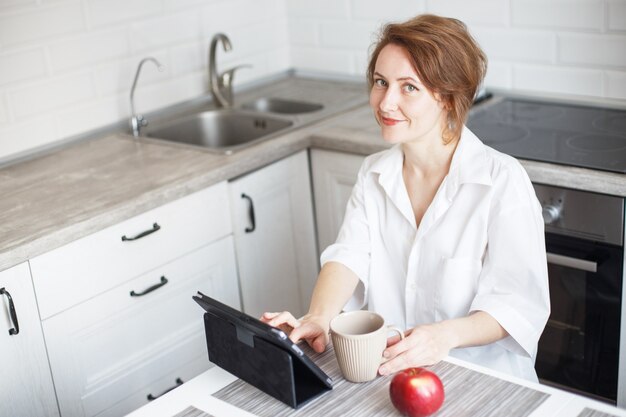 Mujer con taza de café o té usando la computadora portátil en la cocina de casa