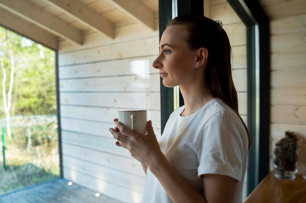 Mujer con taza de café de pie en la sala de estar mirando por la puerta de la terraza