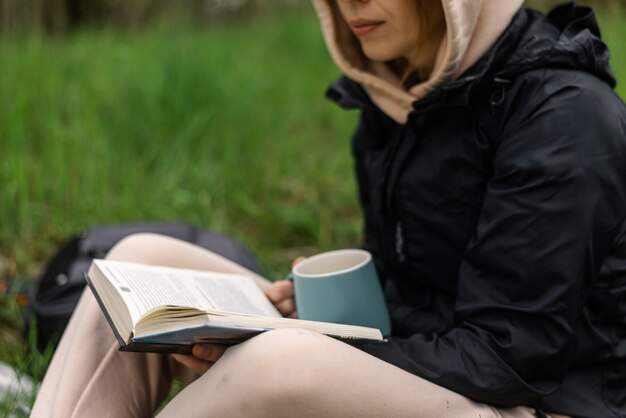 Una mujer con una taza y un libro en el parque se sienta en la hierba verde en un fresco día de otoño.