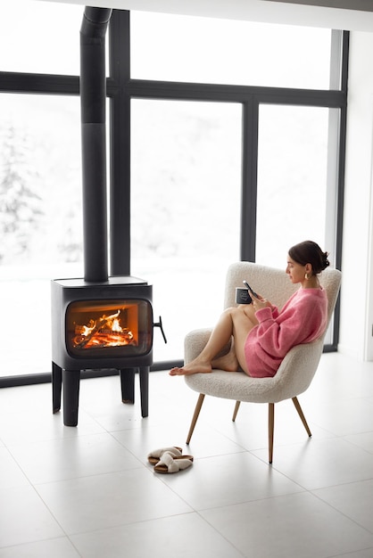 Mujer con taza en silla junto a la chimenea en casa en la naturaleza