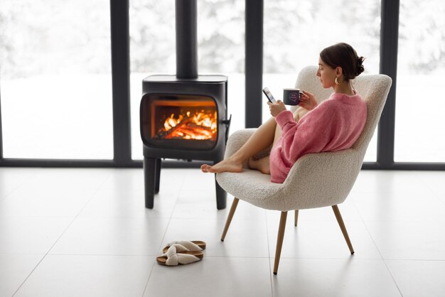 Mujer con taza en silla junto a la chimenea en casa en la naturaleza