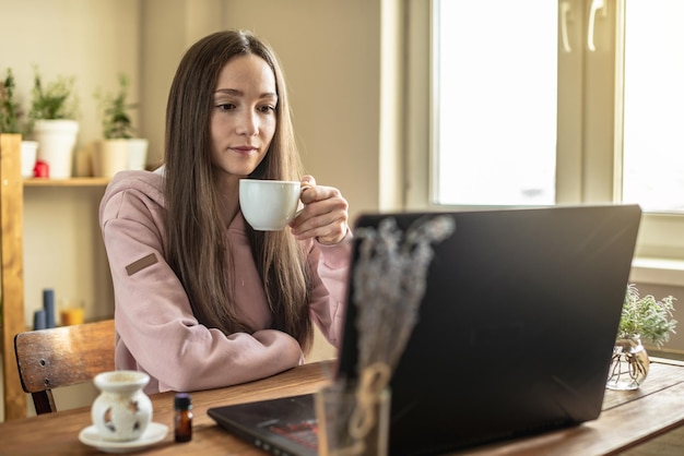Una mujer con una taza de té está sentada frente a la pantalla de una computadora en un ambiente tranquilo y agradable y escucha atentamente el entrenamiento en video o una sesión en línea o simplemente se comunica con un amigo