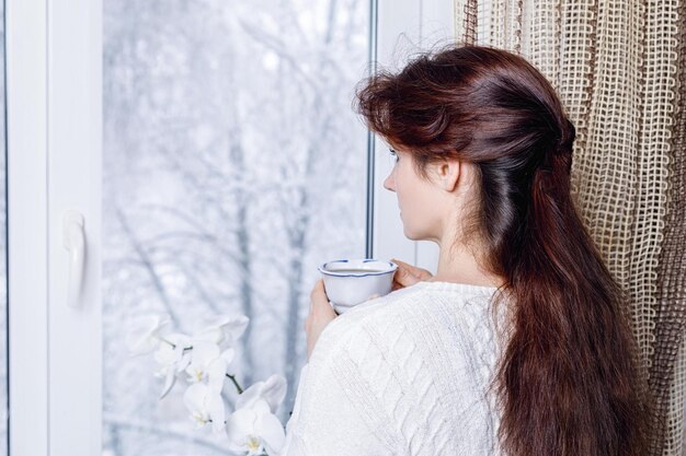 Mujer con taza de té mirando por la ventana con naturaleza cubierta de nieve