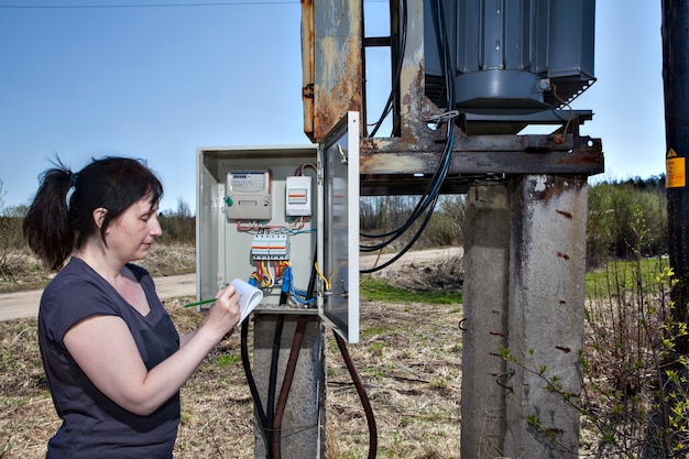 Foto mujer técnico comprobando el medidor de electricidad y la factura de pie cerca de la subestación del transformador de potencia de la aparamenta eléctrica al aire libre.