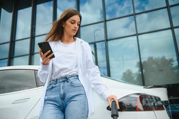 Mujer con teléfono cerca de un coche eléctrico de alquiler Vehículo cargado en la estación de carga