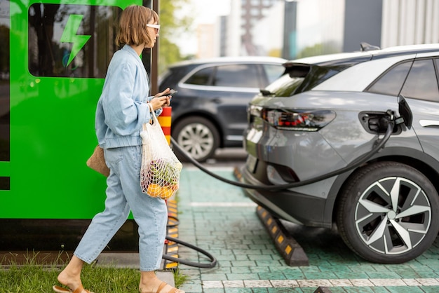 Mujer con un teléfono inteligente y comestibles esperando que su coche eléctrico sea cargado al aire libre