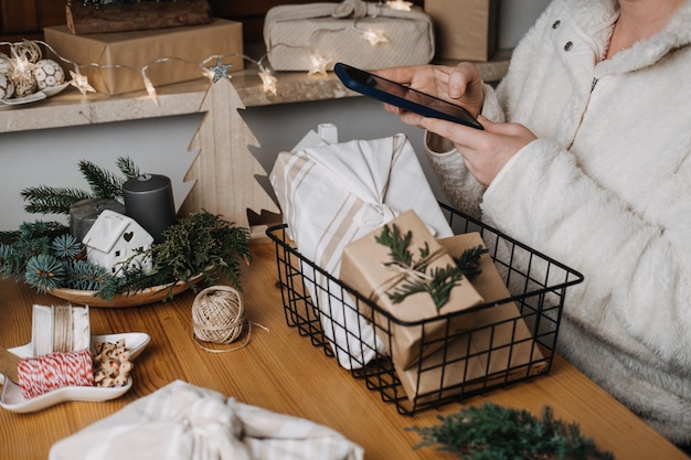 Mujer con teléfono inteligente en la mano y regalos de Navidad ecológicos envolviendo en la mesa Mujer usando teléfono celular cerca de regalos en papel artesanal en la mesa en casa