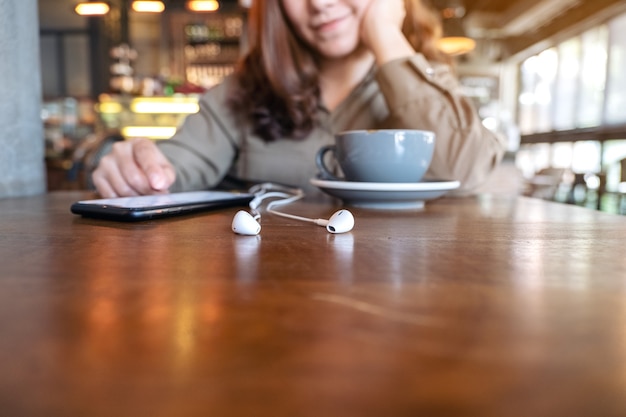 Mujer con teléfono móvil con auriculares y taza de café en la mesa de madera en el café