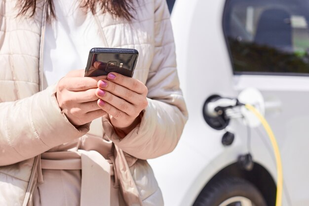 Mujer con un teléfono móvil cerca de recargar coche eléctrico. Carga de vehículos en la estación de carga pública al aire libre. Concepto de coche compartido